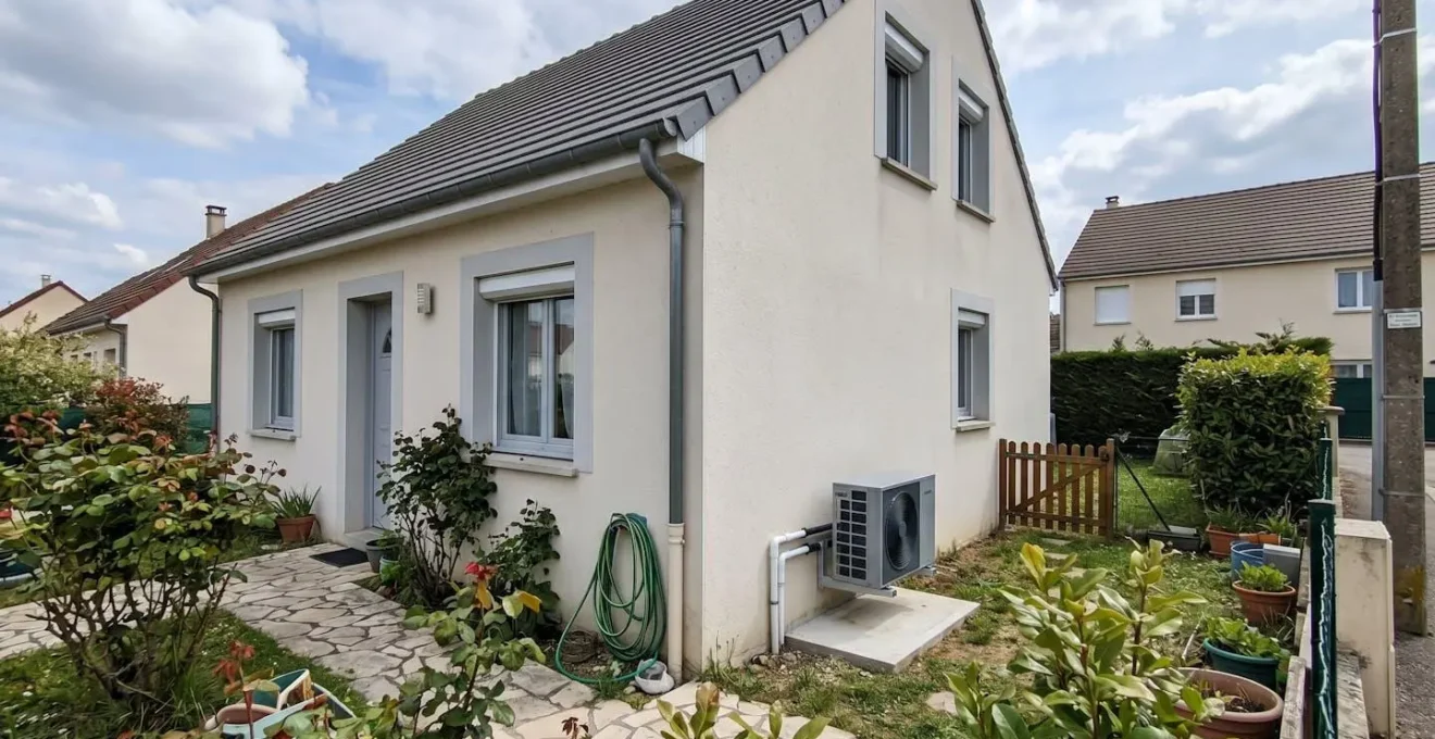 Petite maison contemporaine française avec unité extérieure de pompe à chaleur compacte installée sur le mur latéral, jardin modeste en zone périurbaine